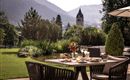 Ein schöner Garten mit einem Tisch, auf dem Frühstück und Getränke serviert werden. Im Hintergrund sind Berge und ein Kirchturm sichtbar.