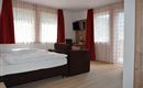 Ein helles Zimmer mit einem Doppelbett und einem gemütlichen Sessel. Große Fenster lassen viel Licht hinein und die roten Vorhänge sorgen für einen warmen Akzent.