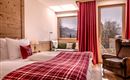 Ein gemütliches Hotelzimmer mit einem Doppelbett und einer rot karierten Decke. Durch das Fenster hat man einen Blick auf die Berge und viel Tageslicht.