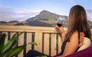 Eine Frau sitzt auf einem Balkon und genießt ein Glas Rotwein. Im Hintergrund ist ein malerischer Berg mit grünen Wiesen zu sehen.
