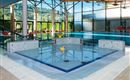 Ein modernes Hallenbad mit hellem Wasser und großen Fenstern. In der Mitte schwimmt ein gelbes Gummienten-Spielzeug.