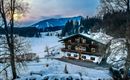 Ein malerisches Alpenhaus im Schnee mit Blick auf die umliegenden Berge. Die Szene wird von einem sanften Sonnenuntergang beleuchtet.