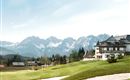 A picturesque landscape with a golf course and an elegant building. Majestic mountains rise in the background under a clear sky.