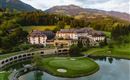 An elegant hotel amidst a green landscape and mountainous backdrop. A golf course and a tranquil lake surround the estate.