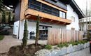 Ein modernes Haus mit einem Holzbalkon und einem gepflegten Vorgarten. Der Außenbereich ist mit Steinen und Pflanzen gestaltet, und es gibt eine Terrasse mit Sitzmöglichkeiten.
