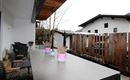 Eine Terrasse mit einem langen Tisch und bunten Töpfen mit Pflanzen. Im Hintergrund sind moderne Häuser und ein Zaun aus Holz zu sehen.