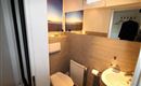 Ein modernes Badezimmer mit einer Toilette und einem Waschbecken. Die Wände sind mit grauen Fliesen und einem Landschaftsbild dekoriert.