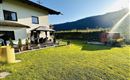 Ein gepflegter Garten mit grünem Rasen und einer Pergola. Im Hintergrund sind Berge und ein schöner blauer Himmel zu sehen.