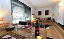 Ein modernes Wohnzimmer mit Holzfußboden und großen Fenstern. Der Esstisch ist festlich gedeckt mit Weinglas und einer Flasche Wein.