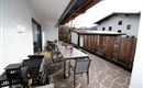 Eine einladende Terrasse mit einem großen Tisch und Stühlen. Hinter der Terrasse ist ein Garten mit einem Holzzaun und einem daneben stehenden Gebäude.