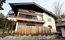 Ein modernes Haus mit einem Holzüberhang und großen Fenstern. Der Eingangsbereich ist von einer Holzlattenzaun und Steinen umgeben.