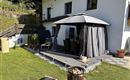 Eine gemütliche Terrasse mit einem grauen Pavillon und Gartenmöbeln. Im Hintergrund steht ein Haus und der Rasen ist gut gepflegt.