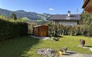 Ein schöner Garten mit einem Holzschuppen und gepflegtem Rasen. Im Hintergrund sind grüne Berge und ein blauer Himmel zu sehen.