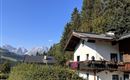 Ein schönes Haus mit Balkon in einer grünen Umgebung. Im Hintergrund sind majestätische Berge und ein klarer blauer Himmel zu sehen.