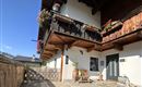 Ein charmantes Haus mit einem Holzbalkon und bunten Blumen. Im Vordergrund gibt es einen kleinen Sitzbereich und viel Tageslicht.