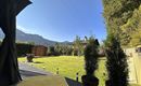 Ein schöner Garten mit grünem Rasen und Bäumen. Im Hintergrund sind Berge und ein strahlend blauer Himmel sichtbar.