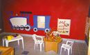 Ein bunter Spielbereich für Kinder mit einem Zug-Motiv an der Wand. Es gibt einen runden Tisch mit Spielzeug und Stühlen.
