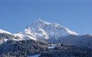 Ein schneebedeckter Berg unter einem klaren blauen Himmel. Im Vordergrund sind bewaldete Hänge zu sehen.