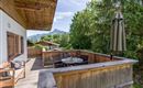 Eine gemütliche Terrasse mit Holzgeländer und einem Tisch. Im Hintergrund sind Bäume und Berge sichtbar.