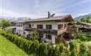 Ein schönes Haus im alpinen Stil mit Balkon und Garten. Im Hintergrund sind grüne Wiesen und Berge zu sehen.