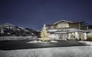 Ein Hotelgebäude im Schnee bei Nacht, umgeben von Bergen. Ein festlich geschmückter Weihnachtsbaum ist im Vordergrund zu sehen.