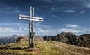 Ein großes, modernes Kreuz steht auf einem grünen Gipfel. Im Hintergrund erstrecken sich majestätische Berge unter einem klaren Himmel.