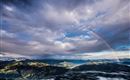 Eine beeindruckende Berglandschaft unter einem bewölkten Himmel mit einem Regenbogen. Die Wolken und die Sonne schaffen ein malerisches Lichtspiel über den Tälern.