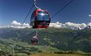 Eine Seilbahn fährt über eine grüne Berglandschaft. Im Hintergrund sind schneebedeckte Berge und ein klarer blauer Himmel zu sehen.