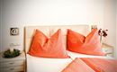Ein gemütliches Schlafzimmer mit einem großen Bett und auffälligen, orangen Kissen. An der Wand sind dekorative Elemente und ein kleines Arrangement von Blumen zu sehen.