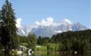 Ein ruhiger See umgeben von grünen Bäumen und beeindruckenden Bergen im Hintergrund. Der Himmel ist klar und blau mit einigen Wolken.