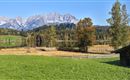 Eine ruhige Landschaft mit grünen Wiesen und bunten Herbstbäumen. Im Hintergrund sind majestätische Berge und ein klarer blauer Himmel zu sehen.