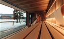Ein modernes Sauna-Innenraum mit Holzregalen und großen Fenstern. Im Hintergrund ist eine winterliche Landschaft mit Schnee und Bäumen zu sehen.