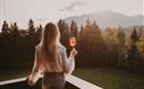Eine Frau steht auf einem Balkon und hält ein Getränk in der Hand. Im Hintergrund sind grüne Wiesen und Berge bei Sonnenuntergang zu sehen.