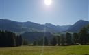 Eine weitläufige grüne Wiese umgeben von Bergen unter einem strahlend blauen Himmel. Die Sonne scheint hell und sorgt für eine schöne, friedliche Atmosphäre.
