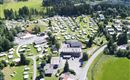 Ein großer Campingplatz mit vielen Wohnmobilen und Stellplätzen in einer grünen Landschaft. Im Hintergrund sind Wälder und sanfte Hügel zu sehen.