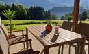 Eine gemütliche Terrasse mit einem Tisch und Stühlen, umgeben von üppiger Natur. Im Hintergrund sind sanfte Hügel und Bäume zu sehen.