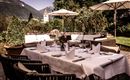 Ein elegantes Esszimmer im Freien mit einem dekorierten Tisch und Blick auf die Berge. Sonnenschirme und Pflanzen umgeben den Bereich und schaffen eine einladende Atmosphäre.