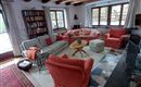 Ein gemütliches Wohnzimmer mit rustikalen Holzbalken und großen Fenstern. Es enthält ein rotes Sofa, einen runden Tisch und einen bunten Teppich.