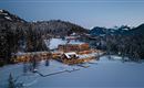 Ein malerisches Winterdorf, umgeben von schneebedeckten Bergen und Tannenbäumen. Die Gebäude sind sanft beleuchtet und reflektieren eine gemütliche Atmosphäre.
