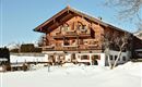 Ein traditionelles alpenländisches Holzhaus in einer schneebedeckten Landschaft. Im Hintergrund sind Berge und ein klarer blauer Himmel sichtbar.