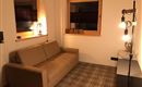 Ein gemütliches Wohnzimmer mit einem beigen Sofa und einem modernen Stehlampe. Es gibt zwei Fenster und die Wände sind hell gestrichen.