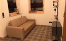 Ein gemütliches Wohnzimmer mit einem beigen Sofa und einem modernen Stehlampe. Die Wände sind hell und es gibt zwei Fenster mit Blick nach draußen.