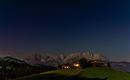 Eine ruhige Nachtlandschaft mit beleuchteten Hütten vor majestätischen Bergen. Der Himmel ist klar und voller Sterne.