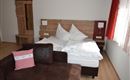 Ein gemütliches Schlafzimmer mit einem Bett, weißen Kissen und einem dunklen Sofa. Helle Wände und Fenster lassen viel Licht herein.