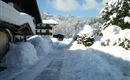 Eine verschneite Straße zwischen traditionellen Häusern. Im Hintergrund sind schneebedeckte Berge zu sehen.